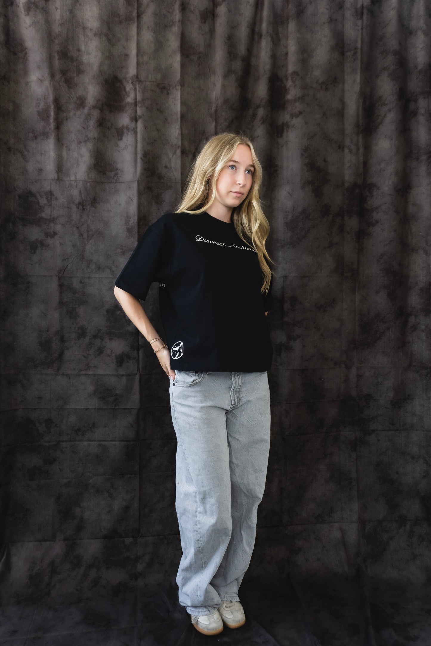 Person wearing a black t-shirt and light gray pants against a dark textured backdrop
