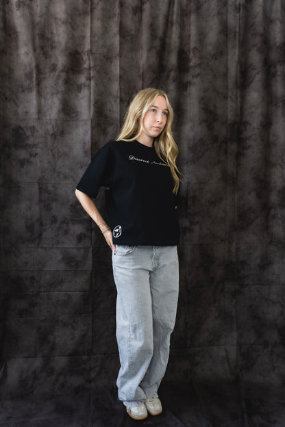 Person wearing a black t-shirt and light gray pants against a dark textured backdrop