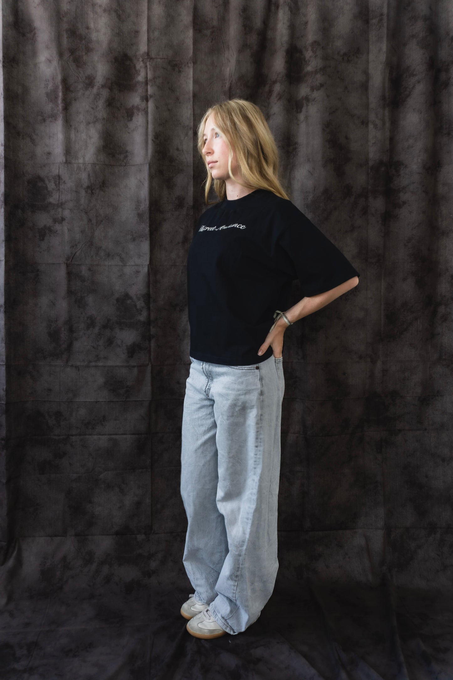 Person wearing a black t-shirt and light gray pants against a dark textured backdrop