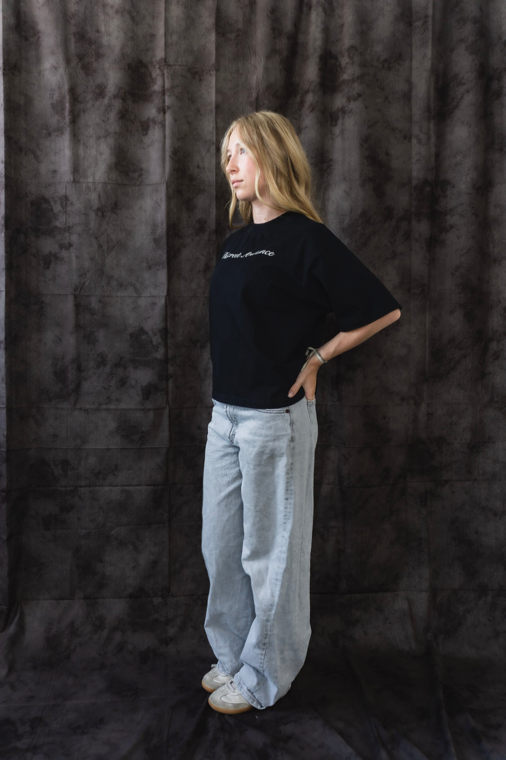 Person wearing a black t-shirt and light gray pants against a dark textured backdrop