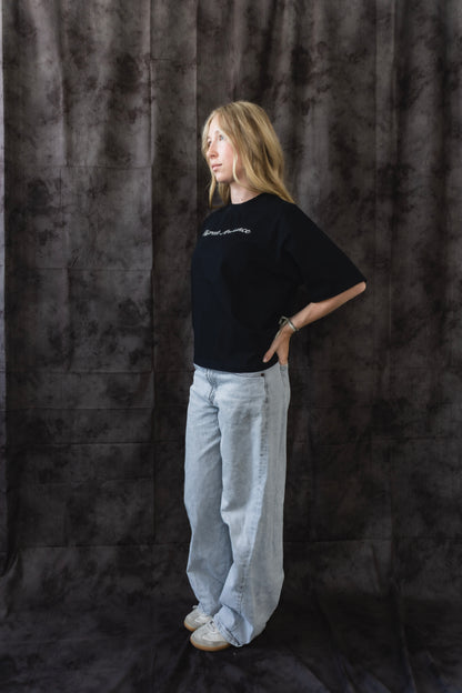 Person wearing a black t-shirt and light gray pants against a dark textured backdrop