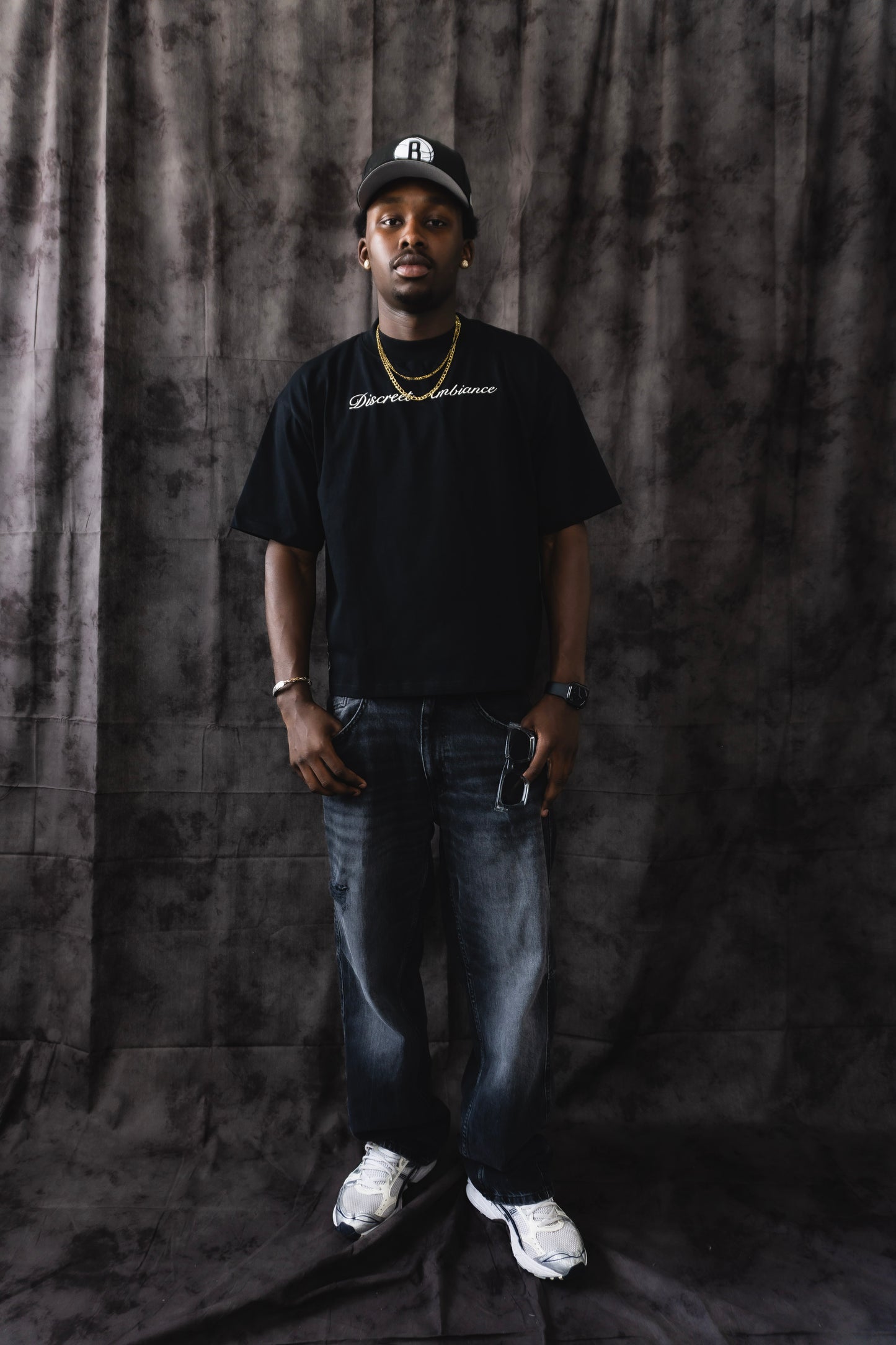 Person wearing a black t-shirt and jeans standing against a dark curtain.