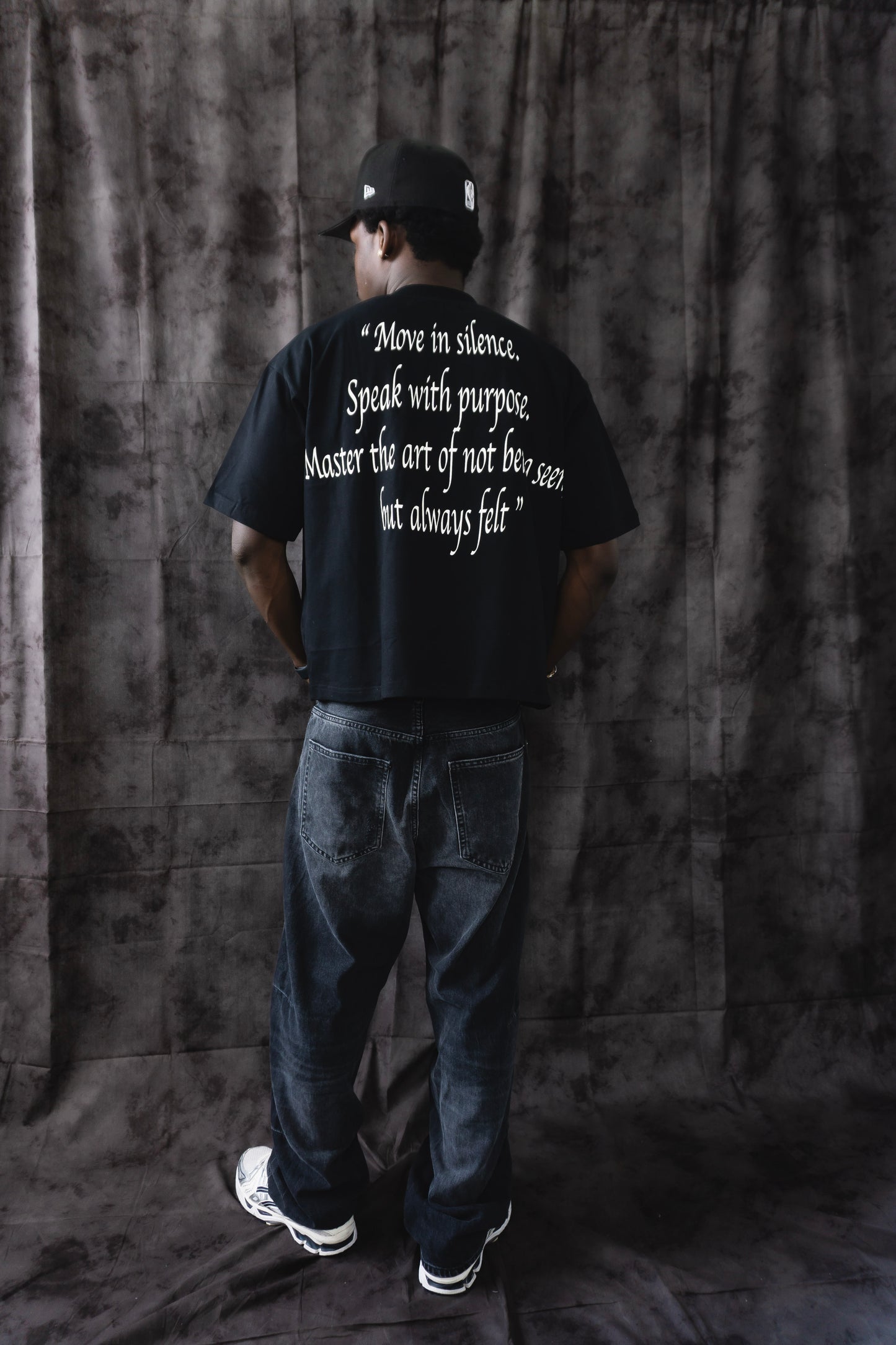 Person wearing a black t-shirt with text on the back, standing against a dark curtain.
